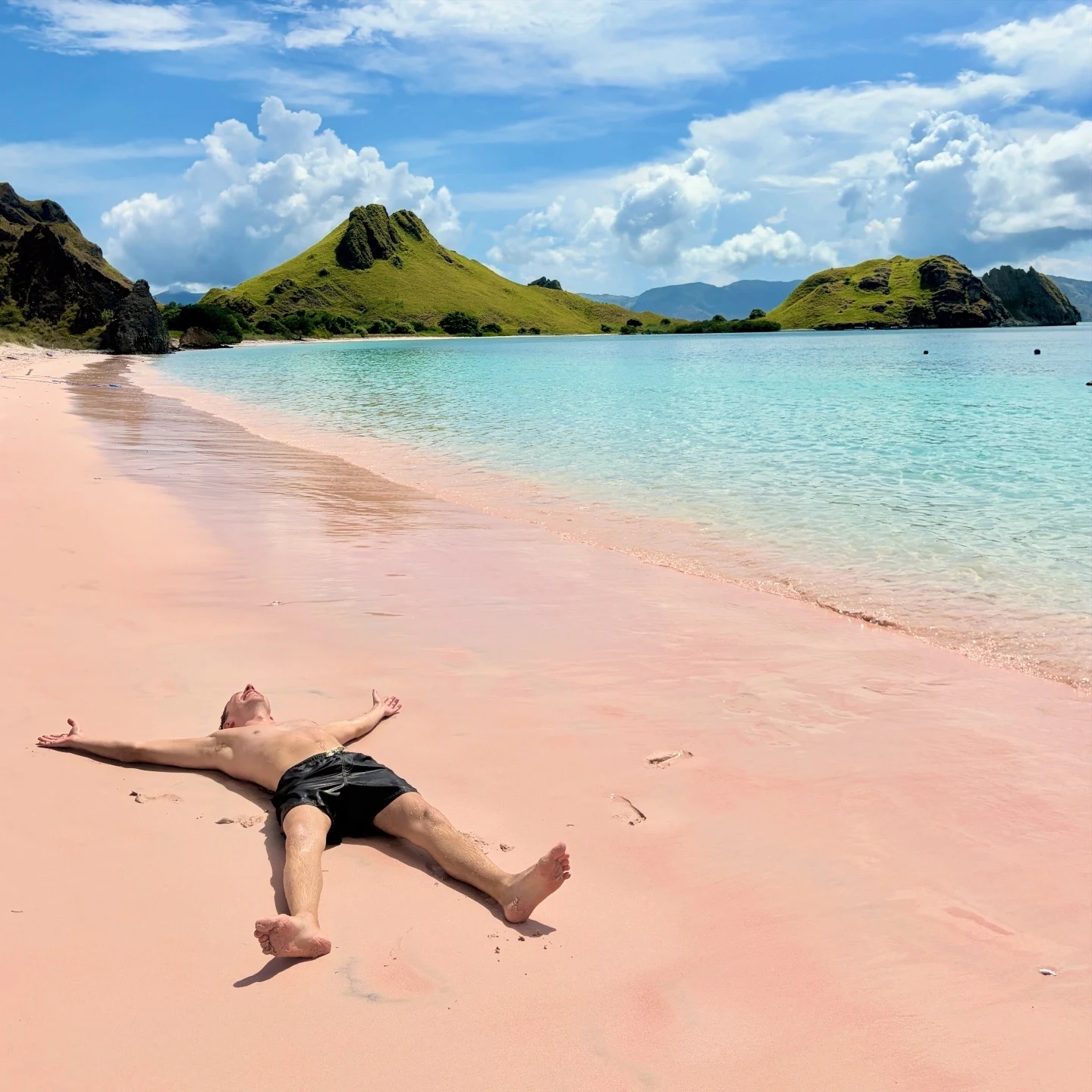 Max lying on a sandy beach with arms and legs outstretched beside turquoise water